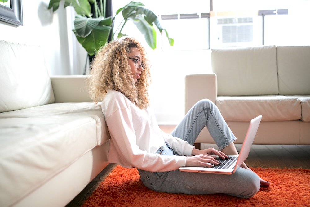 Women with glasses and curly hair sitting on orange rug with an open laptop