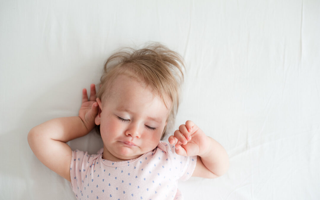 Sleepy baby wearing a pale pink shirt has messy hair and is stretching as they wake from sleep.