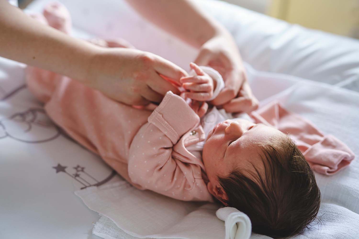 Newborn with dark hair, wearing pink footed pyjamas and laying on a change table is being dressed by an adult whose hands are buttoning the front of the clothes.