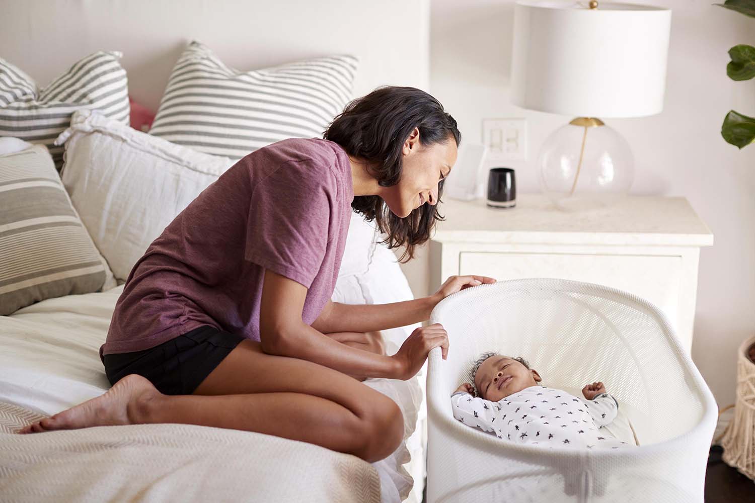 Black woman in a pink shirt and black shorts leans over a sleeping baby in a white bassinet