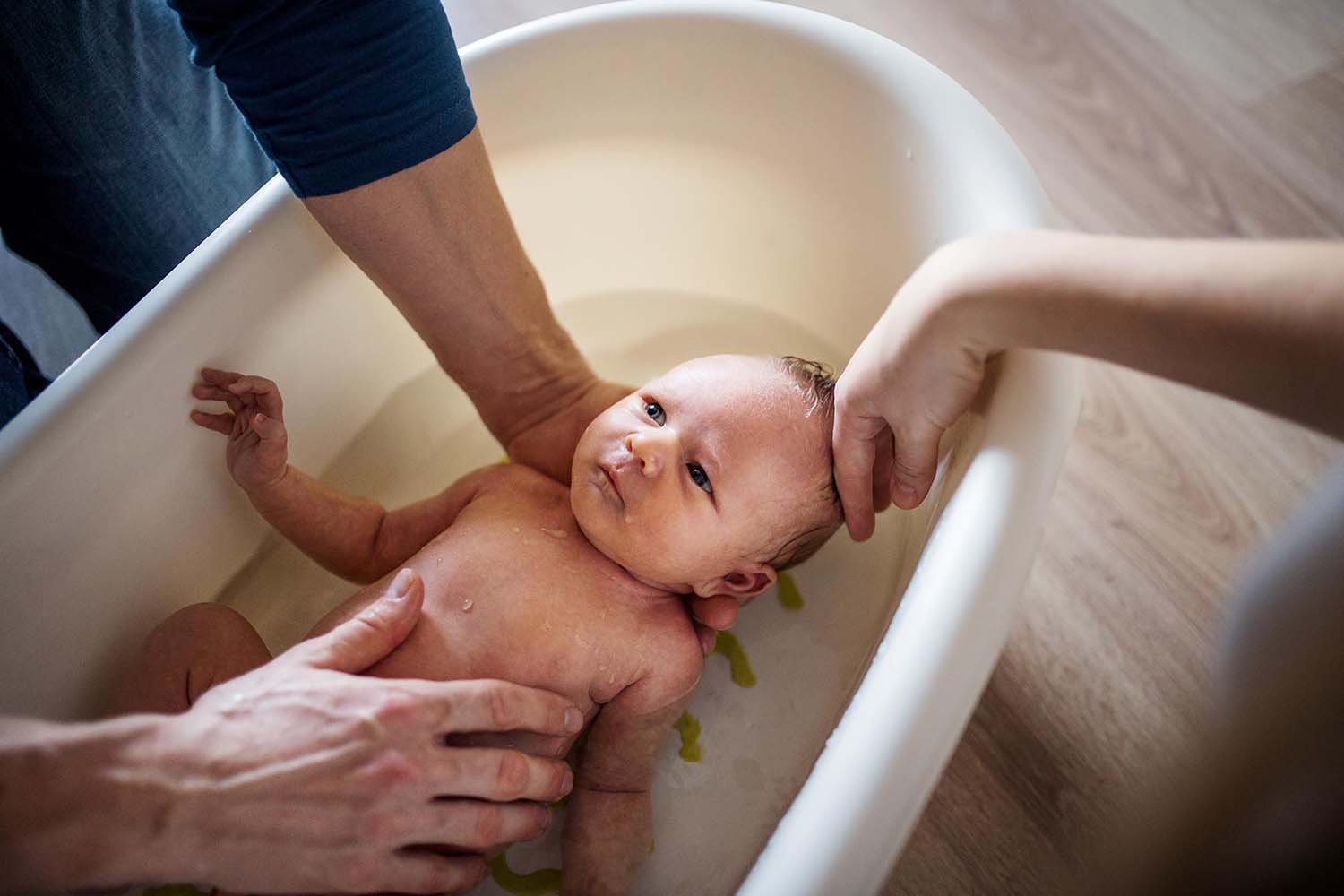 Newborn held by male parent's hands in bath while another hand gently touches baby's head.