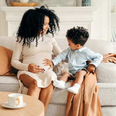 A pregnant mother in a cream coloured dress sits with her toddler who is dressed smartly and touches her belly