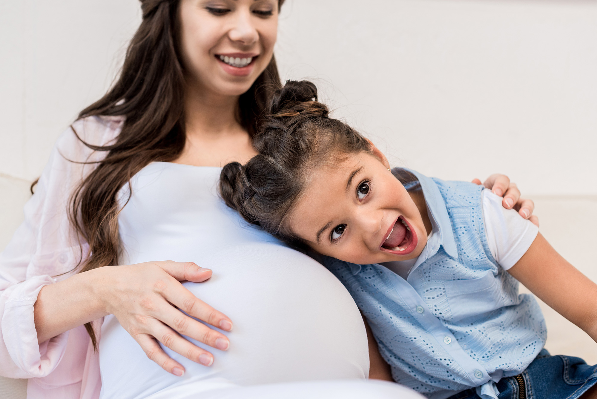 Pregnant woman with long brown hair puts her hand on her pregnant belly as a young girl with a happy face listens to the belly.