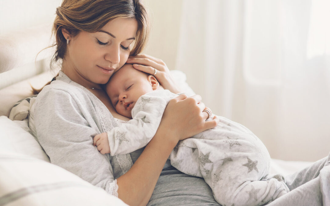 White woman with brown hair wearing a white tshirt has her eyes closed and her cheek on her baby's head as she holds them on her while the baby sleeps.