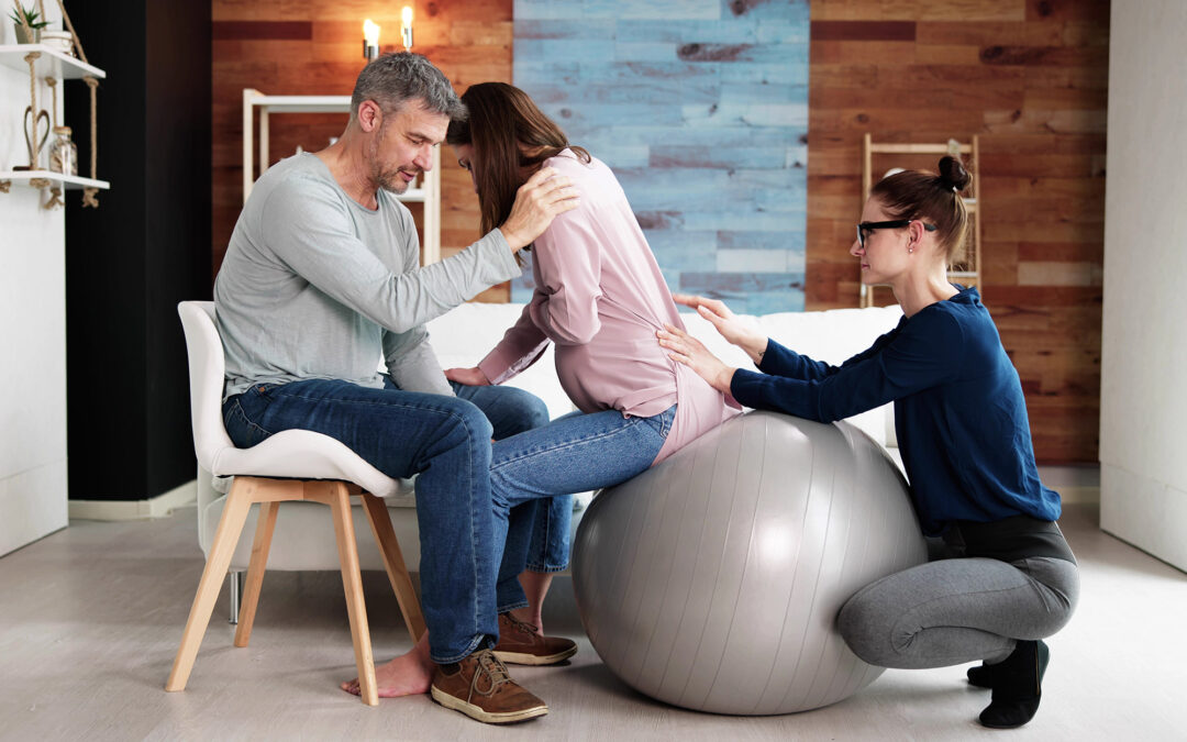 Pregnant woman in jeans and a pink top sits on a grey birth ball and leans into her male partner while a doula with glasses and her hair in a bun, squats behind her and presses into her hips.